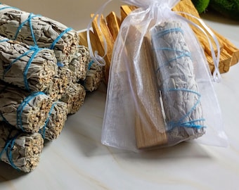 May include: Bundles of white sage, tied with blue twine, are stacked next to a sheer white bag containing a sage bundle and a palo santo stick. The background includes more palo santo sticks.