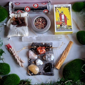 以下が含まれることがあります： ライダータロットデッキ、お香、クリスタル、セレナイトワンド、セージの束など、スピリチュアルなアイテムの詰め合わせ。 大理石の表面に配置され、緑色の苔に囲まれています。