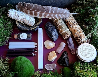 May include: A selection of spiritual items, including sage bundles, crystals, candles, and a feather, arranged on a purple surface and surrounded by green moss. A small jar with a label and a tin of incense cones are also present.