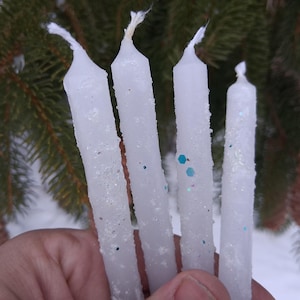 May include: A hand holds four white taper candles with glitter accents. The candles are cylindrical and have a textured, icy appearance. The background features evergreen branches and pine cones, suggesting a winter or holiday theme.
