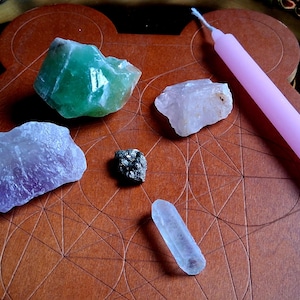 May include: A collection of crystals and a pink candle arranged on a brown wooden surface. The crystals include green, purple, and clear quartz, and a small pyrite stone. The candle is a light pink color.