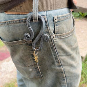 May include: A hand-forged dark grey metal keychain hook with a twisted, scrolled design. The hook is attached to a belt loop on light blue denim jeans and features a small gold-toned metal split ring with a lobster clasp hanging from the bottom.
