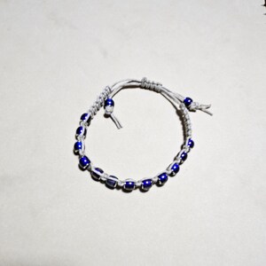 May include: A white cord bracelet with blue beads. The bracelet is tied with a knot and has a small white bead at the end of the cord.