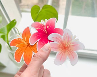 Puede incluir: Tres pegatinas de flores, una rosa, una naranja y una melocotón. Las pegatinas están sobre un fondo blanco.