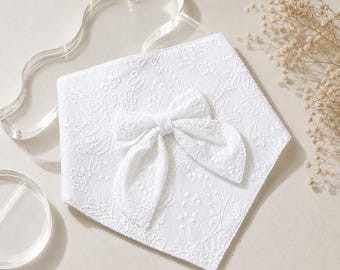 May include: A white triangular bandana featuring an intricate tone-on-tone floral embroidery pattern across the entire fabric surface. A matching white fabric bow is securely attached to the center of the bandana, adding texture and dimension.
