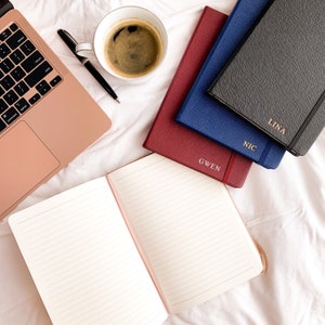 May include: An open lined notebook with a pink spine lies next to a stack of personalized journals in red, blue, and gray. A rose gold laptop, a cup of coffee, and a pen are also on the white surface.