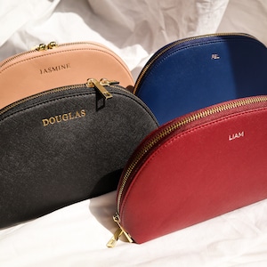 May include: Four half-moon shaped zippered pouches in different colors: black, pink, red, and blue. Each pouch has a gold zipper and a name printed on the front: Douglas, Jasmine, Liam, and Fel.