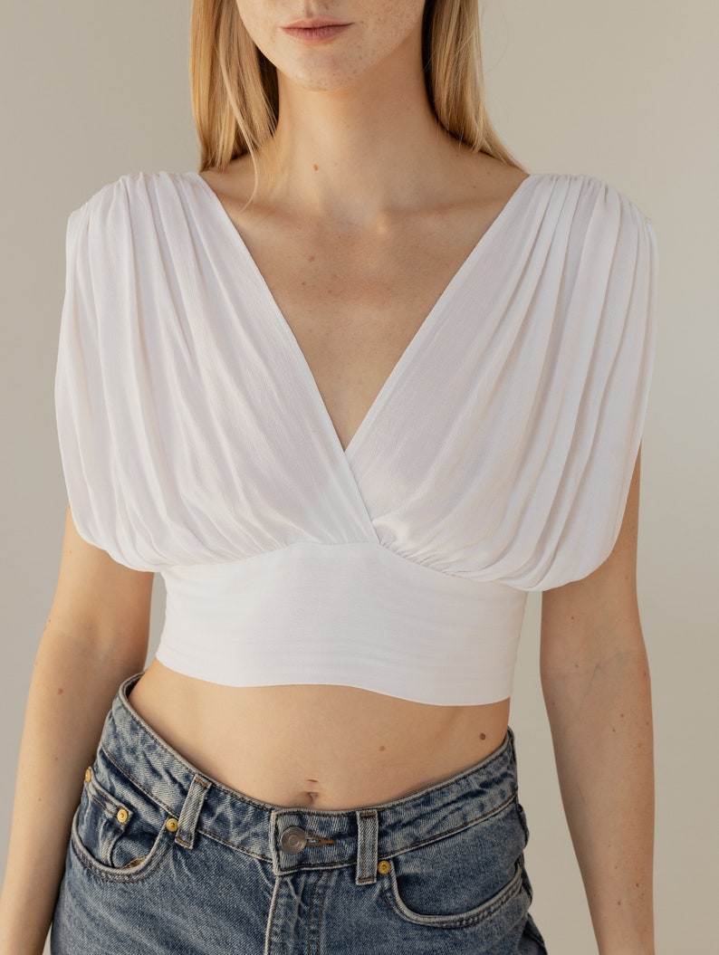 May include: A white crop top with a V-neck and puffed sleeves. The top is made of a soft, lightweight fabric and has a fitted bodice.