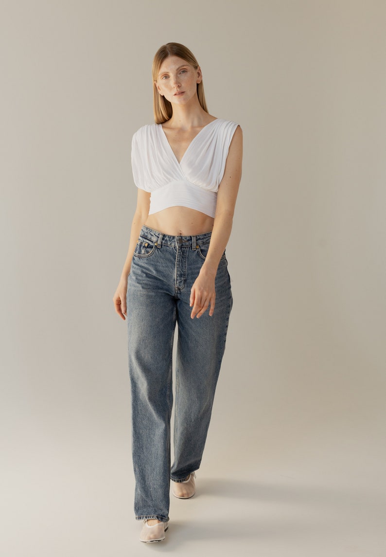 May include: A woman wearing a white crop top with a V-neck and a pair of blue denim jeans.
