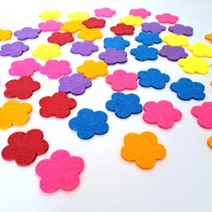 May include: A collection of colorful felt flower shapes in various shades of red, pink, orange, yellow, purple, and blue. The flowers are scattered on a white surface, creating a cheerful and decorative display. These are ideal for craft projects.