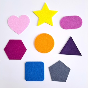 May include: A collection of colorful felt shapes, including a pink heart, yellow star, purple oval, magenta hexagon, orange circle, purple triangle, blue square, and gray pentagon. These shapes are arranged on a white background.