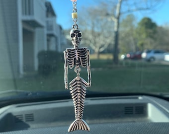 May include: A silver skeleton mermaid car charm hanging from a rearview mirror. The charm has a beaded chain with blue, purple, gray, pink, and yellow beads.