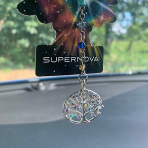 Op de afbeelding: Een zilveren levensboom-hanger met iriserende kristallen hangt aan een zilveren ketting met gekleurde kralen. De hanger is bevestigd aan een kaart met de tekst "SUPERNOVA".