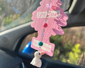Pode incluir: Um ambientador Little Trees rosa em forma de árvore, pendurado no espelho retrovisor de um carro. O ambientador tem as palavras "Little Trees" impressas. Uma pequena figura de coelho branco pende por baixo, presa por uma corrente com contas.