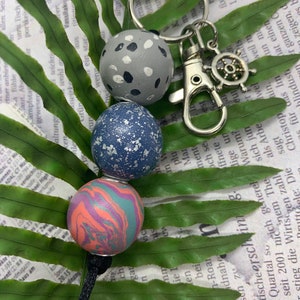 Może przedstawiać: Brelok do kluczy z trzema ozdobnymi koralikami: szarym z białymi kropkami, niebieskim z białymi plamkami oraz pomarańczowo-zielonym marmurkowym wzorem. Brelok ma srebrne zapięcie i zawieszkę w kształcie koła sterowego.