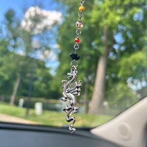 May include: A silver dragon charm hangs from a beaded chain with gray, yellow, red, and black beads. The dragon is detailed with intricate scales and wings. The charm is suspended in front of a blurred green background.