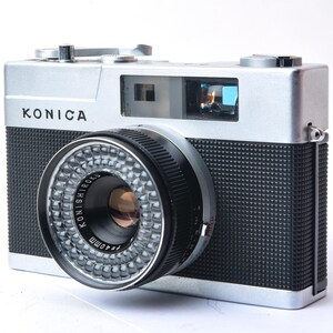 May include: A silver Konica camera with a black lens and a black textured body. The camera has a silver top plate with the word "KONICA" printed on it.