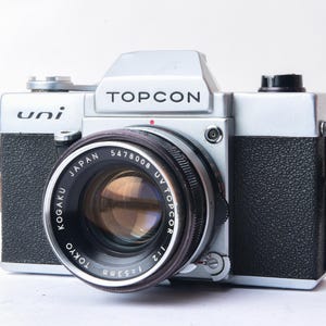 Può includere: Una macchina fotografica a pellicola da 35 mm di colore argento e nero con un obiettivo argentato. La macchina fotografica ha il testo "uni" e "TOPCOR" sul davanti. L'obiettivo è un obiettivo "TOPCOR 1:1.8 5478008 UV" prodotto da "KOGAKU TOKYO JAPAN".
