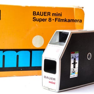 May include: A vintage Bauer Mini Super 8 film camera with its original box. The camera is black and silver with a textured finish, and the box is orange with blue and white text. The camera has a lens and various control buttons.