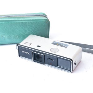 May include: A vintage Minolta camera with a silver body, black accents, and a gray wrist strap. A teal green leather case with the Minolta logo is next to the camera. The camera has a lens, dials, and a viewfinder.