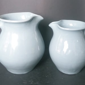 May include: Two light blue ceramic pitchers of different sizes. The larger pitcher has a wider base and a slightly flared lip. The smaller pitcher has a more rounded base and a narrower lip.
