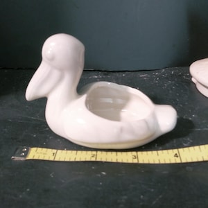 May include: A white ceramic pelican-shaped planter. The pelican has a detailed beak and a hollowed-out area for planting small succulents or flowers. A yellow measuring tape is next to the planter, showing a length of approximately 4 inches.