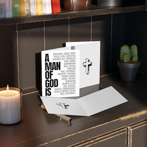 Könnte beinhalten: Eine weiße Grußkarte mit einem schwarzen Kreuz und dem Text "A Man of God Is", gefolgt von einer Liste von Tugenden. Eine brennende Kerze und ein kleiner Kaktus im Topf sind ebenfalls zu sehen.