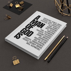 Puede incluir: Un diario blanco con el texto "A Man of God Is" en letras negras en negrita. El diario está rodeado de clips dorados, lápices y un objeto geométrico dorado decorativo sobre una superficie gris oscuro.