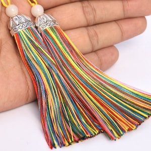 May include: Two colorful tassels with a silver-colored cap and a white bead. The tassels are made of multiple strands of thread in various colors, including red, yellow, green, and blue. The tassels are held in a hand.