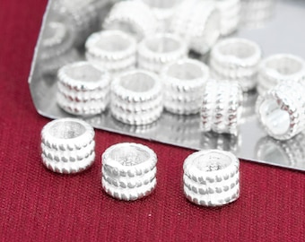 May include: A close-up of a group of silver-coloured metal beads. The beads are round and have a textured surface. They are arranged in a pile on a silver surface.