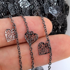 Puede incluir: Collar de cadena negra con tres delicados colgantes en forma de corazón. Cada colgante presenta un diseño de filigrana con un motivo de rosa.