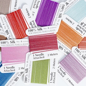 May include: A collection of colorful silk threads for stringing pearls and beads. The spools are in various colors, including red, purple, pink, green, and orange. Each spool is labeled with the text "100% Silk" and "2 Meters".