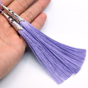 May include: Two lavender silk tassels with silver metal tops. The tassels are hanging from a hand.