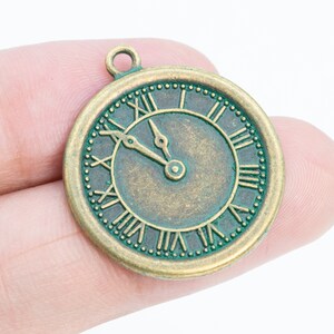 Puede incluir: Un dije de reloj de metal verde y dorado con marcas de números romanos. Las manecillas del reloj están configuradas a las 12:00.