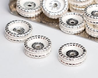 Può includere: Perle in metallo color argento con un bordo rialzato e testurizzato e un piccolo foro al centro. Le perle sono rivestite di smalto bianco con un centro nero.