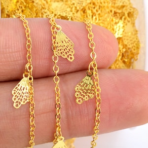 May include: Gold chain with delicate, ornate, fan-shaped charms. The chain is shown draped over a hand.