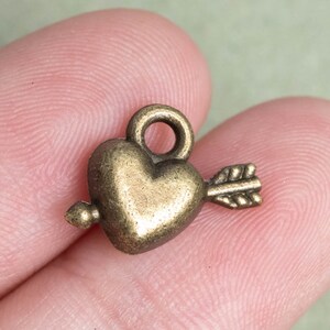 May include: A gold-toned metal charm in the shape of a heart with an arrow through it. The charm has a loop at the top for attaching to a necklace or bracelet.