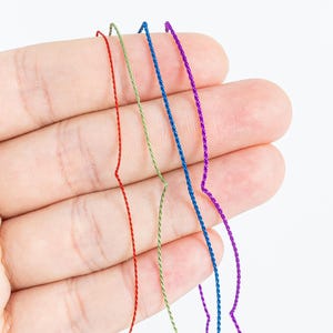 May include: Close-up of four strands of colorful thread: red, green, blue, and purple. The threads are thin and appear to be made of a shiny material. The threads are held in a hand against a white background.