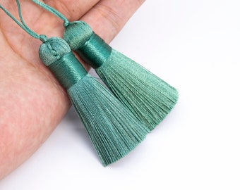 Puede incluir: Dos borlas de seda verde con un nudo en la parte superior y una larga franja. Las borlas se sostienen en una mano.