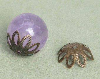 Può includere: Una perla sferica di pietra preziosa viola chiaro con un cappuccio decorativo in metallo color bronzo. Il cappuccio ha la forma di un fiore con petali traforati. La perla e il cappuccio sono esposti su una superficie verde chiaro.
