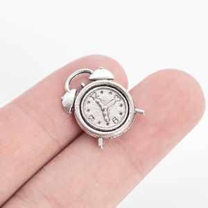May include: Silver-toned alarm clock charm with a white face and black hands. The clock is about 1 inch in diameter.