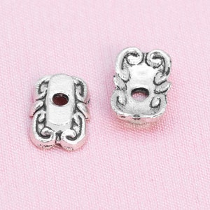 May include: Two silver metal beads with ornate designs and a hole in the center. The beads are on a pink surface.