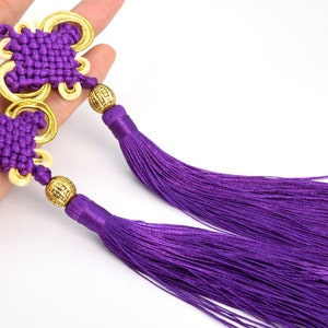 Puede incluir: Dos amuletos de nudo chino de color morado con detalles dorados y borlas. Los nudos tienen forma de mariposa y están atados con un cordón decorativo.