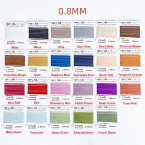 May include: A collection of silk thread spools in various colors, including white, black, gray, and shades of blue, pink, and green. Each spool is labeled with the color name and specifications, including 0.8mm thickness and 2 meters of thread.