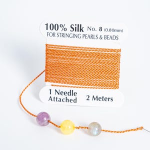 May include: A spool of orange 100% silk thread for stringing pearls and beads. The label reads "1 Needle Attached 2 Meters". Three colorful beads are strung on the thread: purple, yellow, and gray.