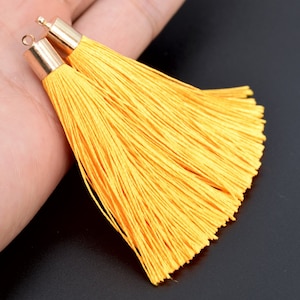 Könnte beinhalten: Zwei gelbe Quasten mit goldenen Metallkappen. Die Quasten bestehen aus weichen, seidenartigen Fäden und sind perfekt, um jedem Projekt einen Hauch von Eleganz zu verleihen.