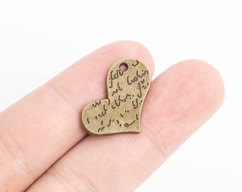 May include: A bronze heart-shaped charm with Arabic script engraved on the surface. The charm has a hole at the top for attaching to a necklace or bracelet.