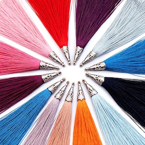 May include: A collection of twelve long, silky tassels arranged in a circular pattern. Each tassel features a vibrant thread color, including pink, red, purple, silver, blue, black, orange, and beige, topped with a decorative silver metal cap.