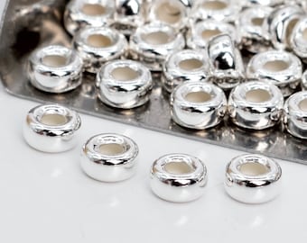 Può includere: Primo piano di perline di metallo color argento con una finitura liscia e lucida. Le perline sono rotonde con un foro al centro. Le perline sono disposte in un mucchio e su una superficie bianca.