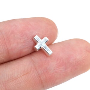 May include: A small, silver-colored cross. The cross is a simple, minimalist design with a slightly rounded shape. The image is a close-up shot, highlighting the details of the cross. The cross is likely a religious symbol, possibly for jewelry or crafts.